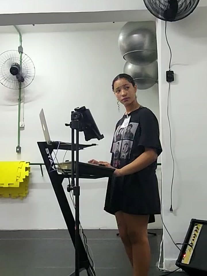 Candidata cantando ao microfone, com o número de inscrição na camiseta, na sala de canto da prova de bolsa