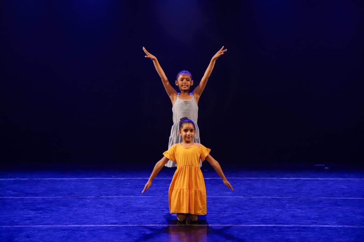 Duas alunas, uma de vestido amarelo, com os braços abertos no palco